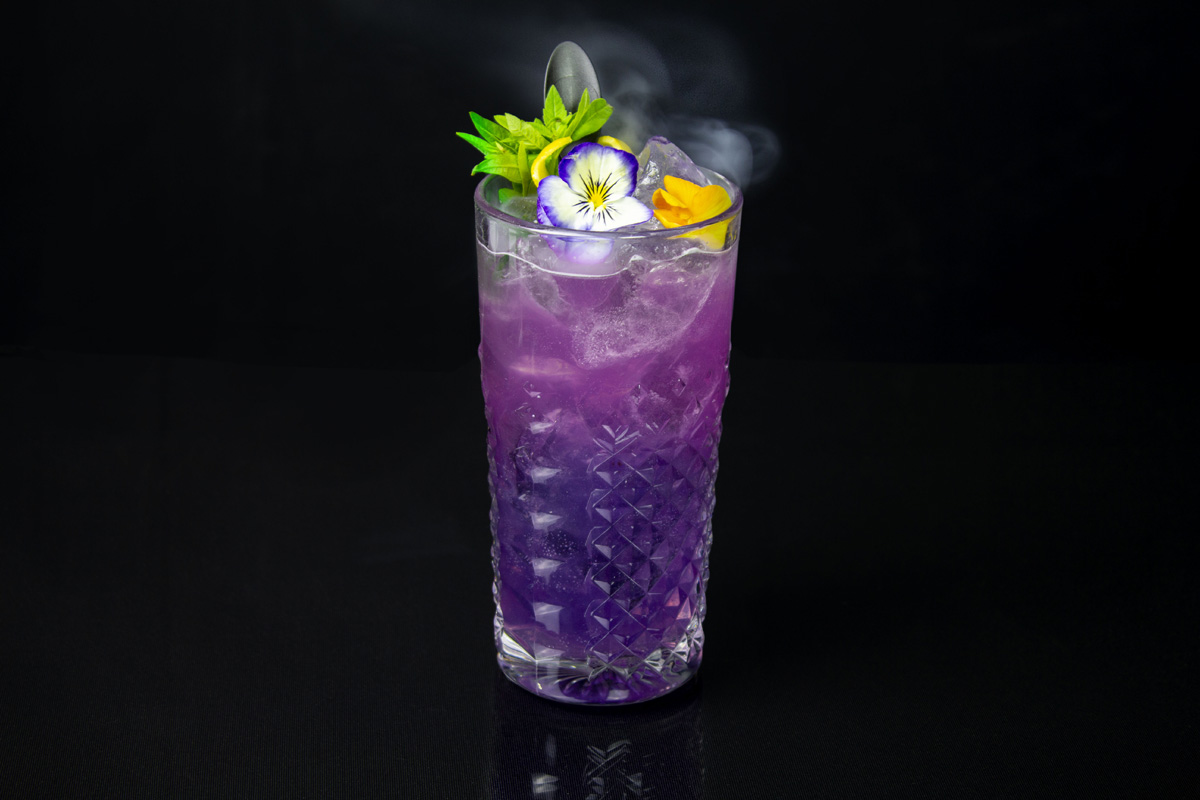 Smoke and Mirrors purple cocktail in a large tumbler topped with viola flowers