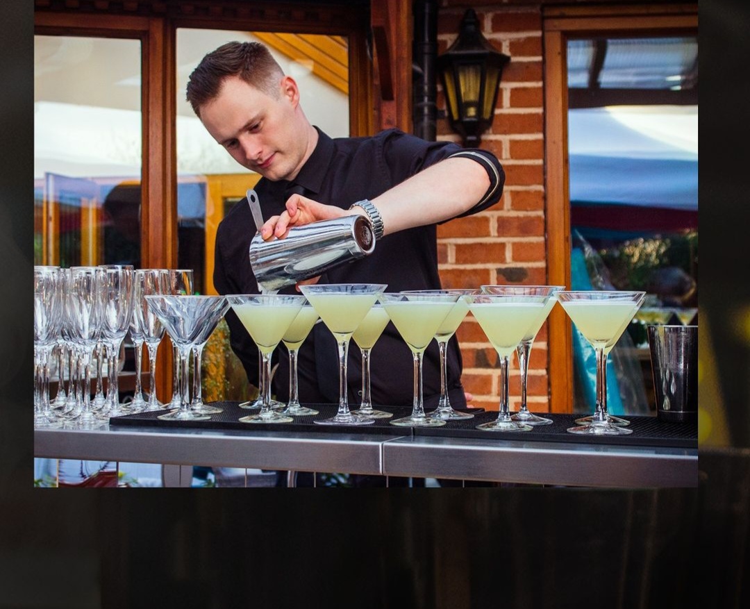 A young male bartender in a black shirt behind a bar, pouring a row of drinks from a cocktail shakers in to martini glasses.