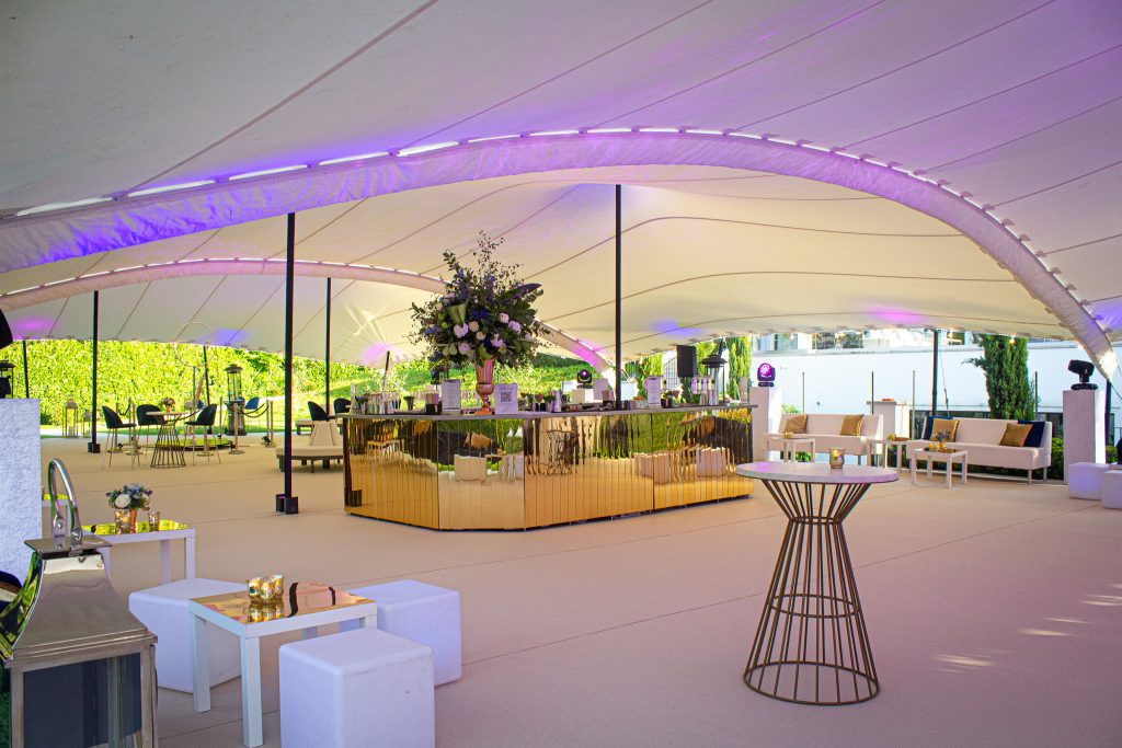 A gold tone centrepiece mirrored bar, surrounded by white casual furniture and poseur tables in a white marquee